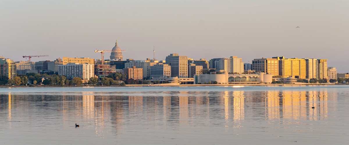 City skyline over Lake Monona, featuring the Wisconsin State Capitol and Monona Terrace. Several construction cranes are visible.